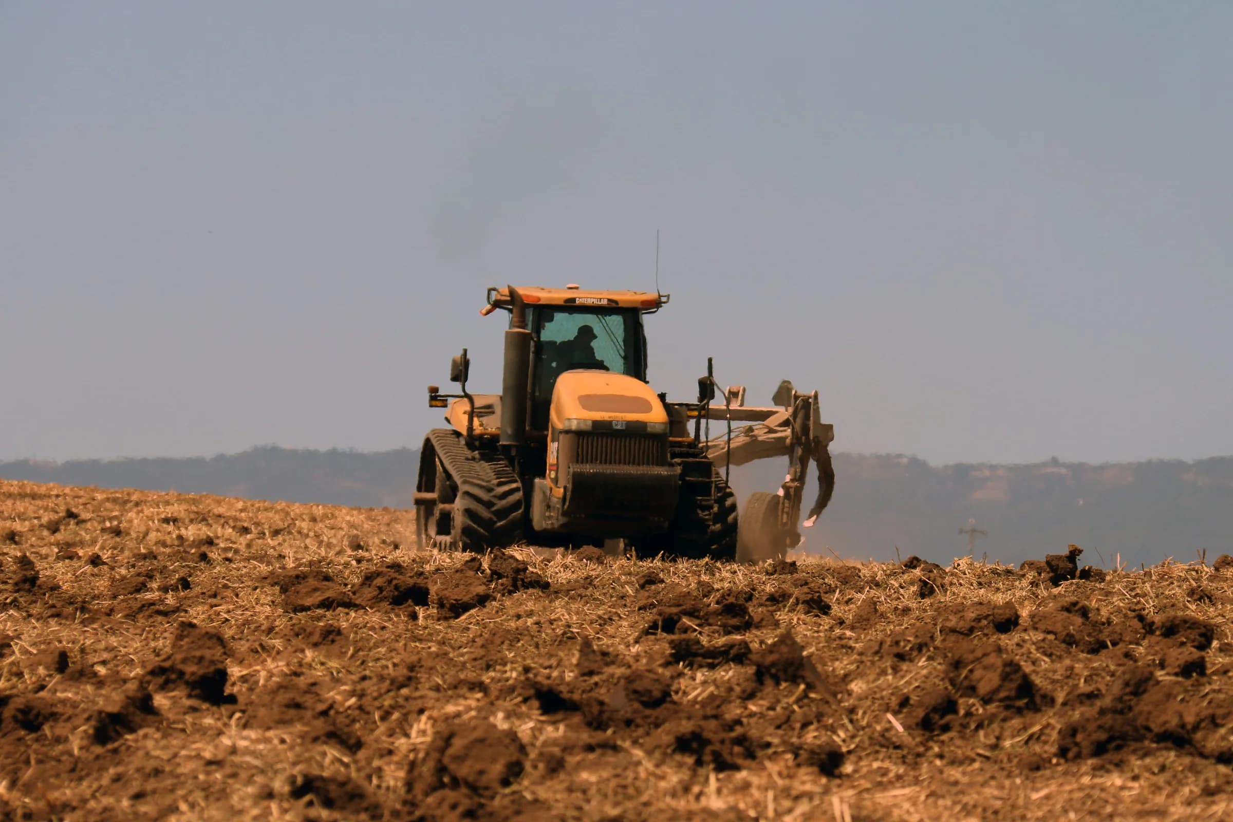 A tracked tractor pulling a tillage implement across a ploughed field, with low hills on the horizon