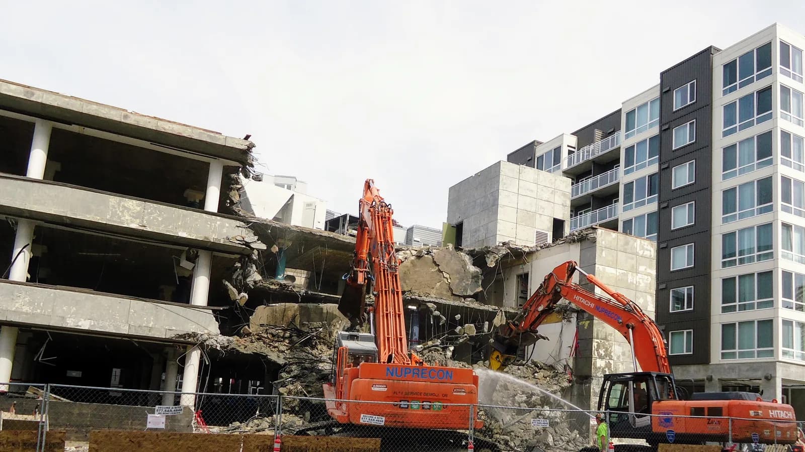An orange excavator demolishing a concrete building beside an apartment block